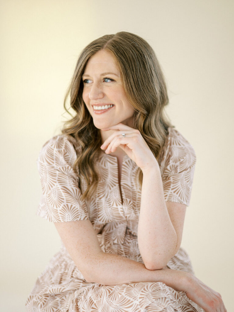 Smiling woman with long wavy hair in a patterned dress, seated with hand under chin, looking to the side against a soft neutral background.
