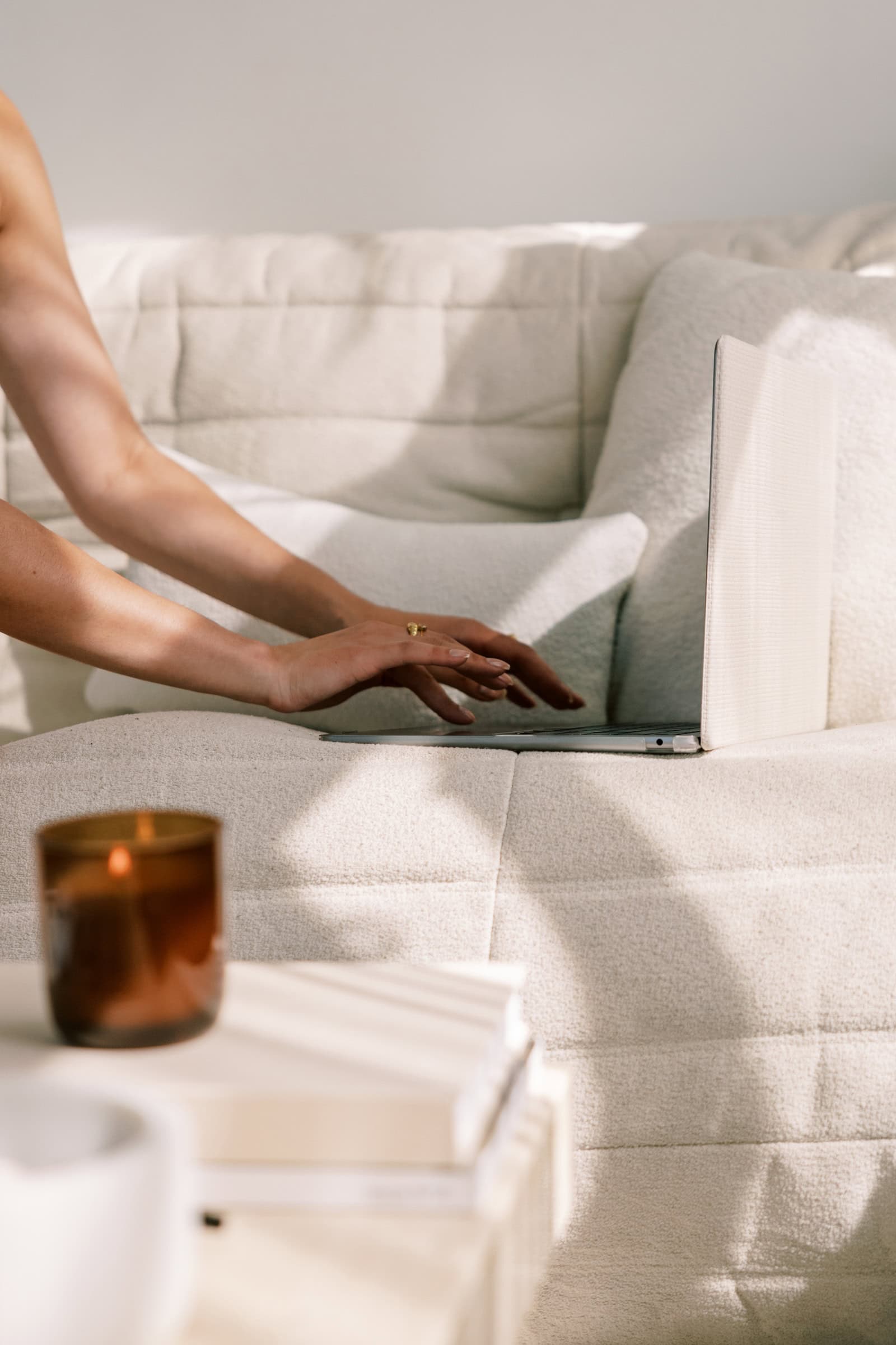 Woman typing on laptop computer in aesthetic space with cream colored couch and amber candle lit