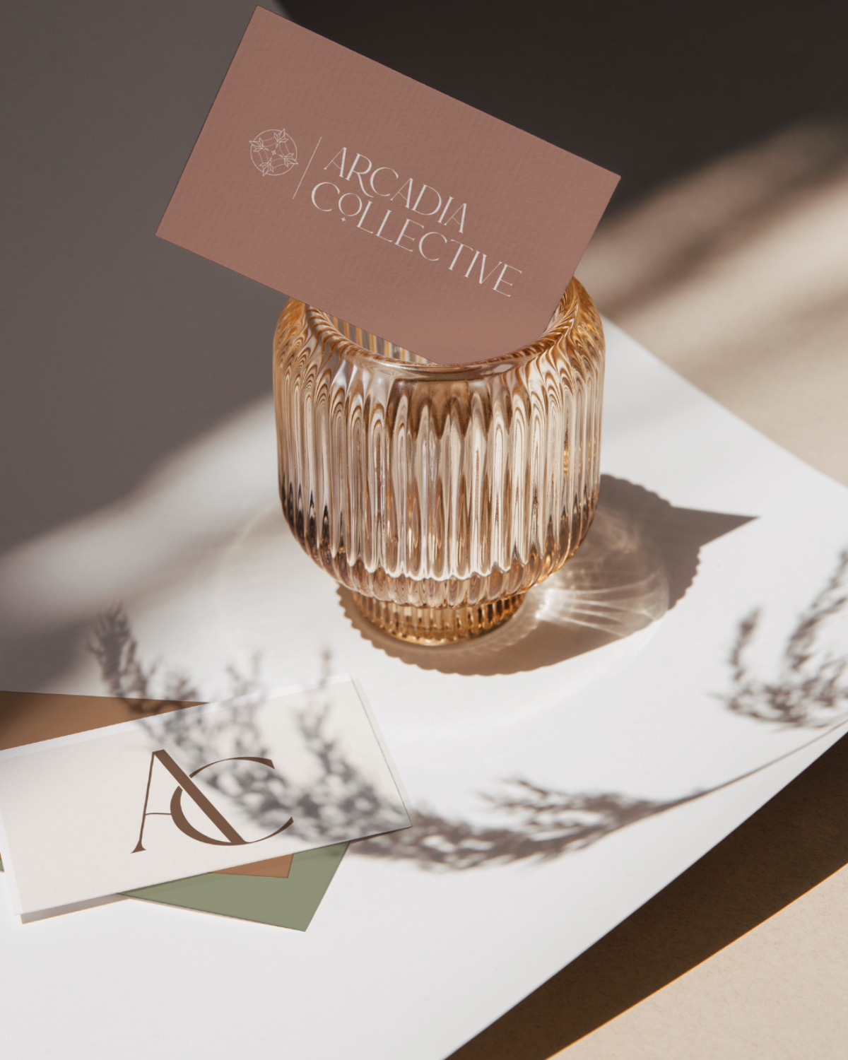 Elegant flat lay of Arcadia Collective business cards in warm terracotta, cream, and sage tones styled in a ribbed glass vase with soft natural light and shadows.