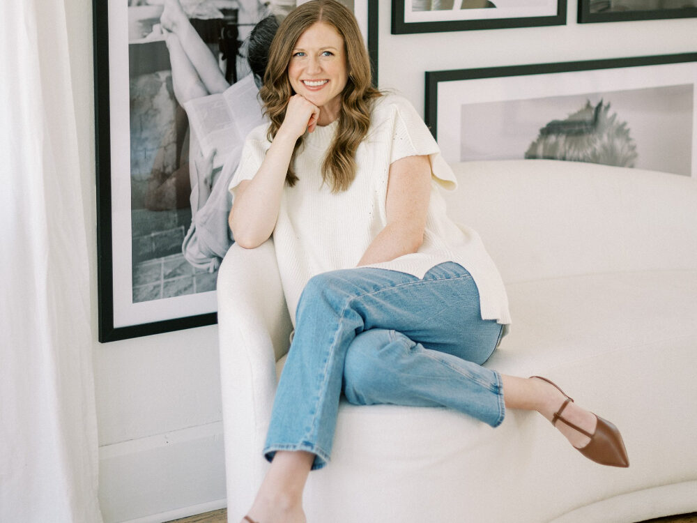 A woman sits relaxed on a white curved sofa, smiling with her hand on her chin. She wears a white top, jeans, and brown heels. Framed black-and-white photos and botanical prints hang behind her.