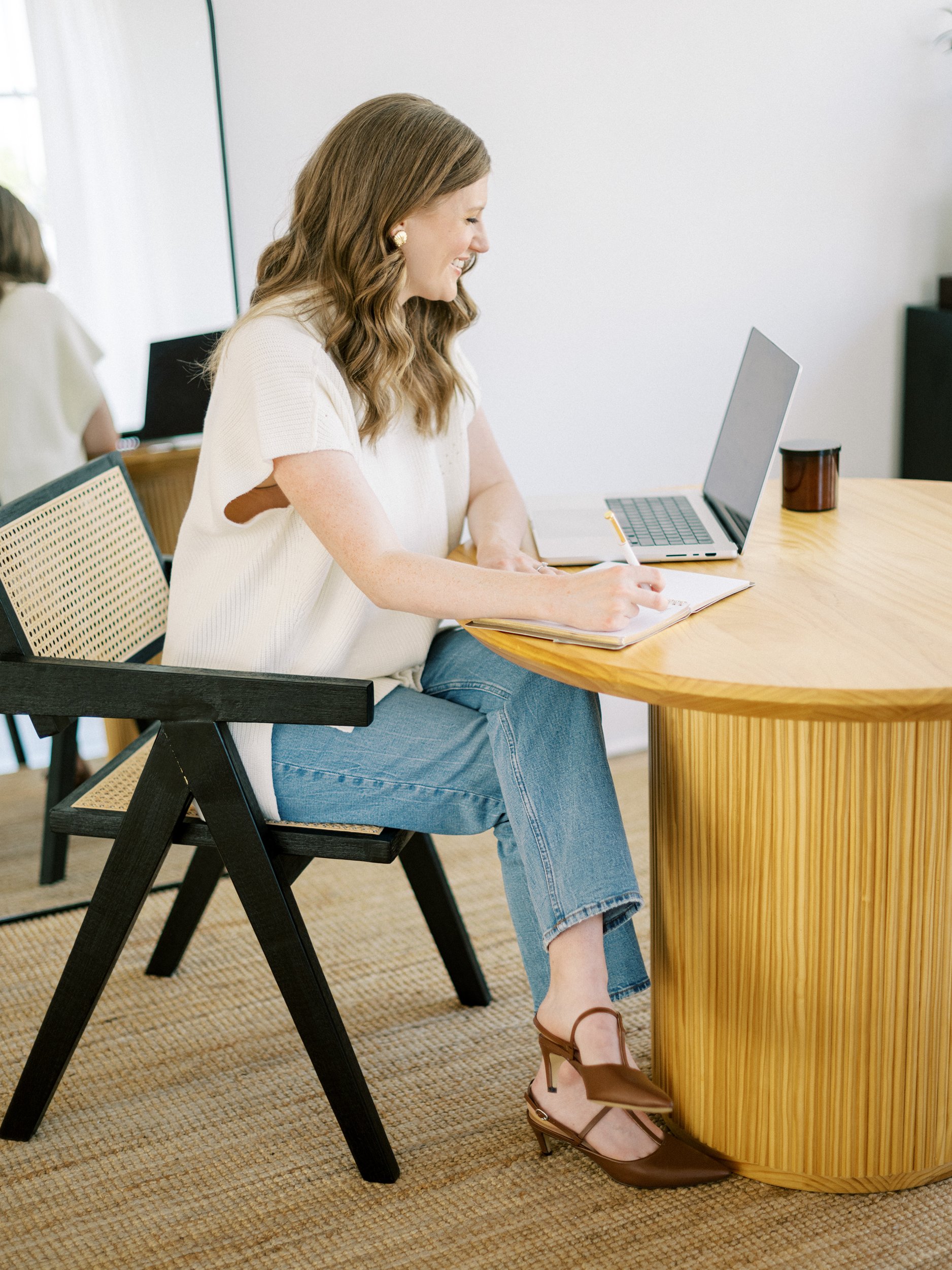 A woman sits at a wooden desk, smiling while writing in a notebook and working on a laptop. She wears a white top, jeans, and brown heels in a bright, modern workspace.