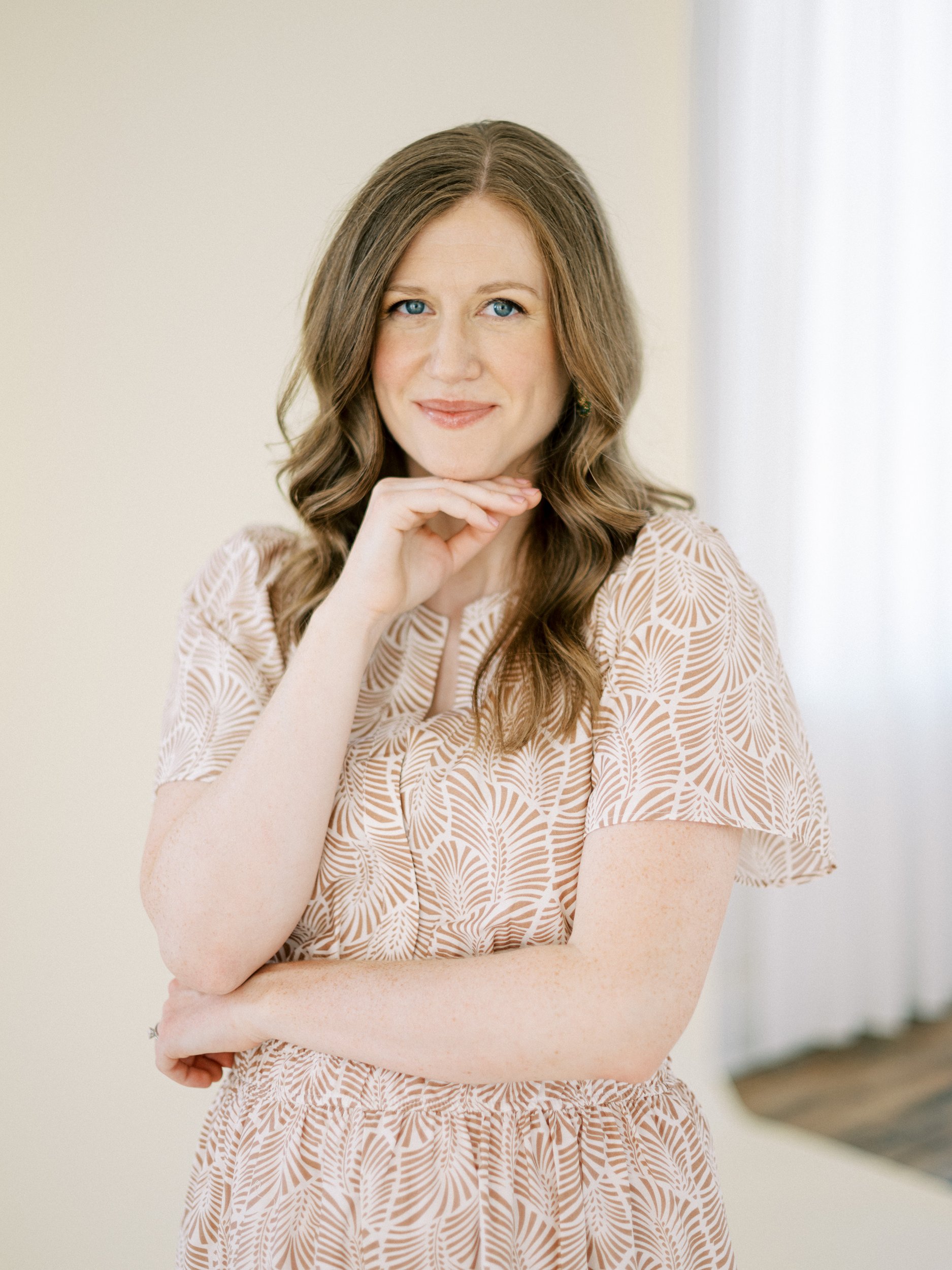 A woman in a beige patterned dress poses with a gentle smile, one arm crossed and the other resting near her chin, standing in a softly lit room with a neutral backdrop.