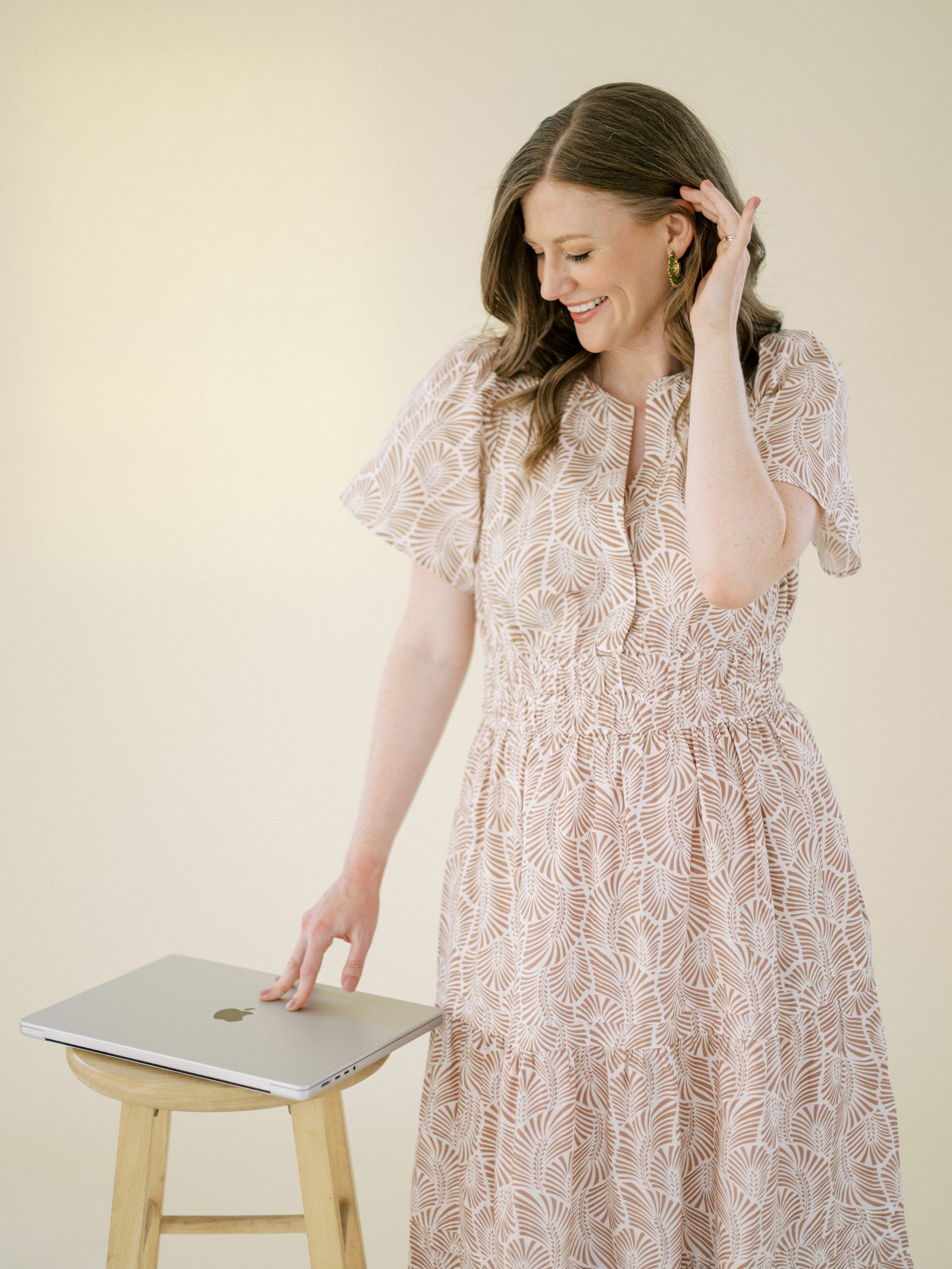 A woman in a patterned beige dress smiles while touching her hair with one hand and resting the other on a closed laptop placed on a wooden stool against a soft cream background.