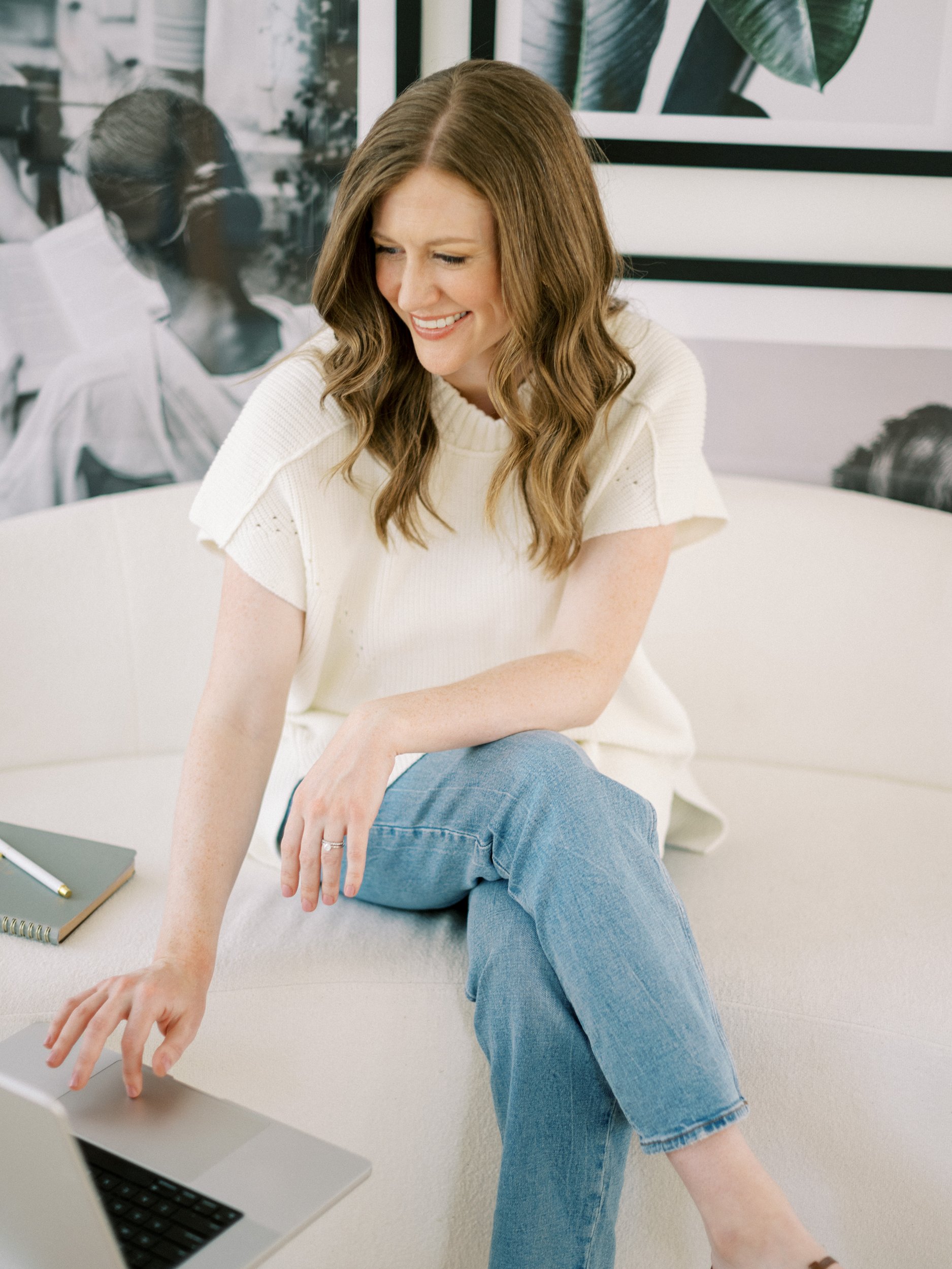Woman sitting on a light sofa, smiling while working on a laptop. She wears a cream knit top and jeans, with framed black and white artwork in the background.