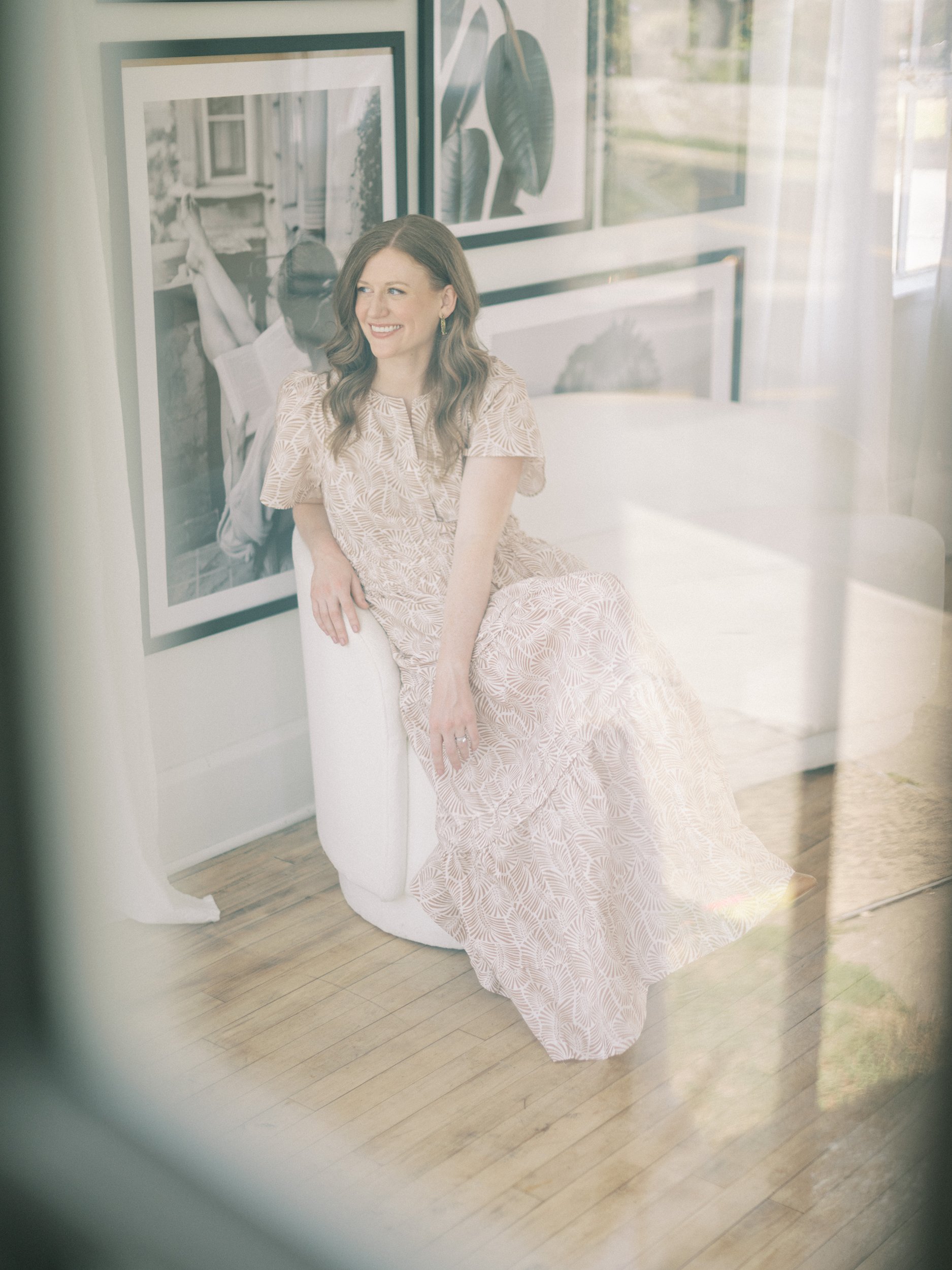 Woman in a long beige patterned dress sitting on a white chair near large framed photos. Soft window reflections create a light, airy feel in the room. | showit review