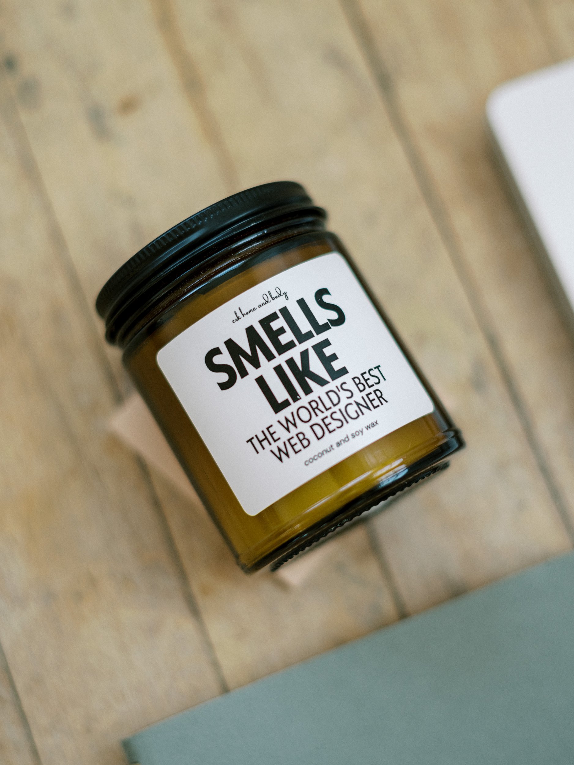 Candle labeled “Smells Like the World’s Best Web Designer” sitting on a wooden surface near a notebook. The candle jar is amber with a black lid. | my honest showit review