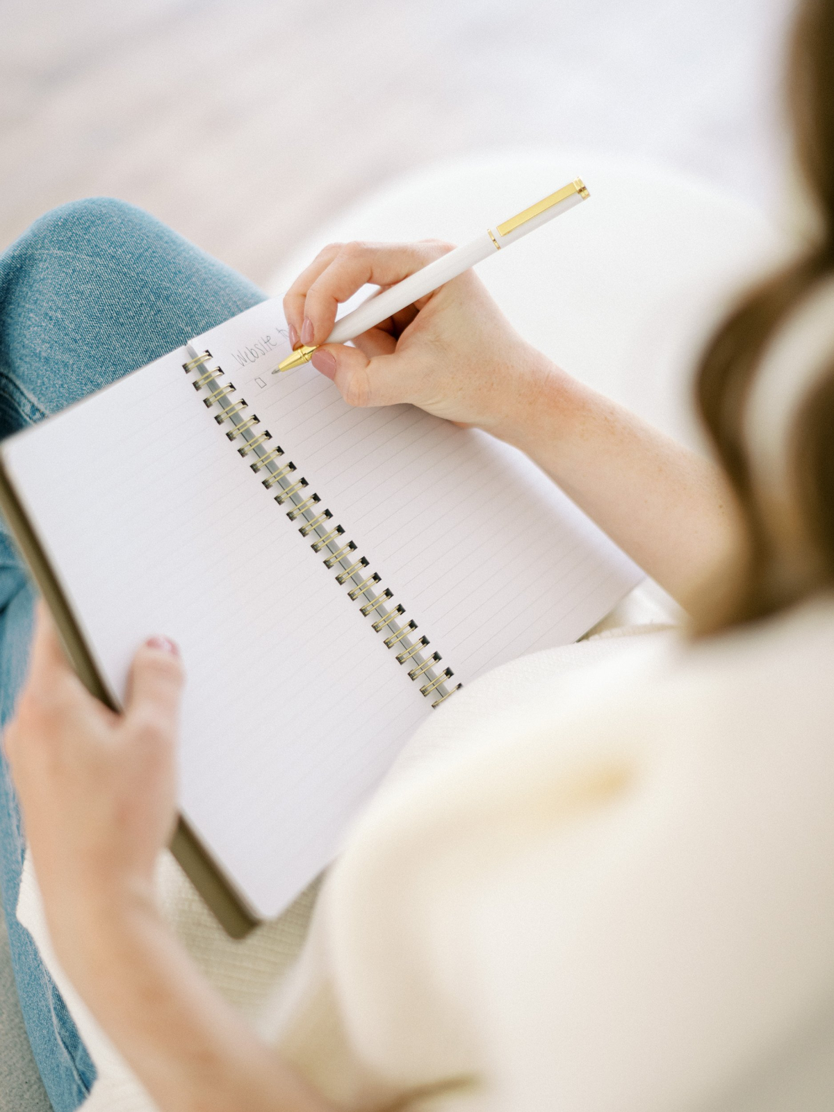 Hand holding a white pen while writing in a spiral notebook, resting on denim jeans in a bright, softly lit setting. | template vs custom website