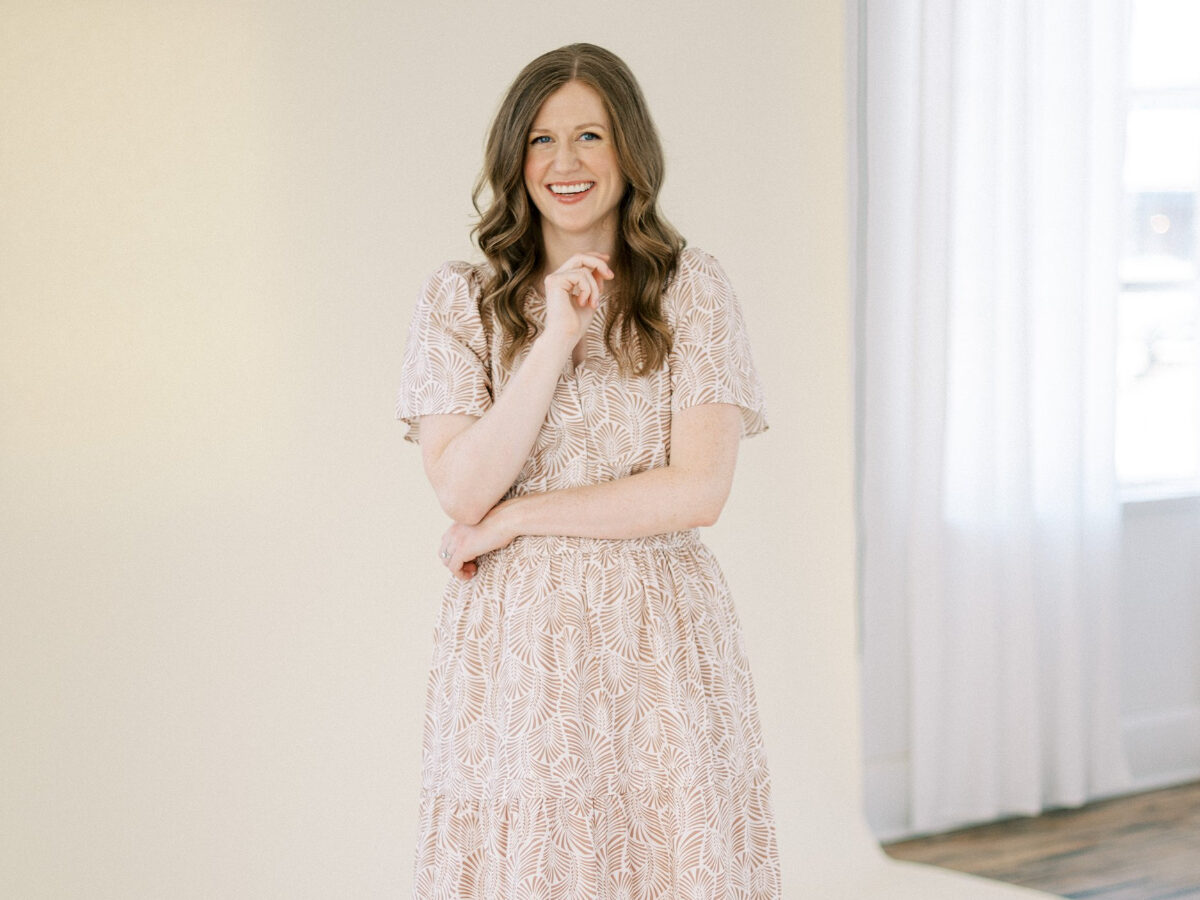Woman standing in a neutral studio space wearing a light patterned dress, smiling confidently with arms crossed.