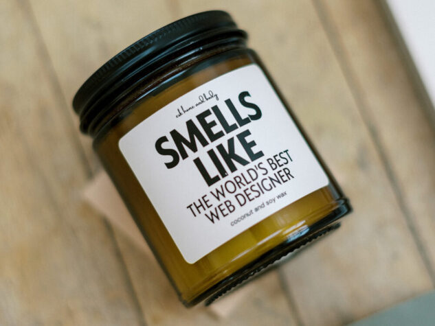 Amber glass candle jar on a wooden surface with a label reading “Smells Like the World’s Best Web Designer,” styled as a playful, branded desk accessory. | showit pricing