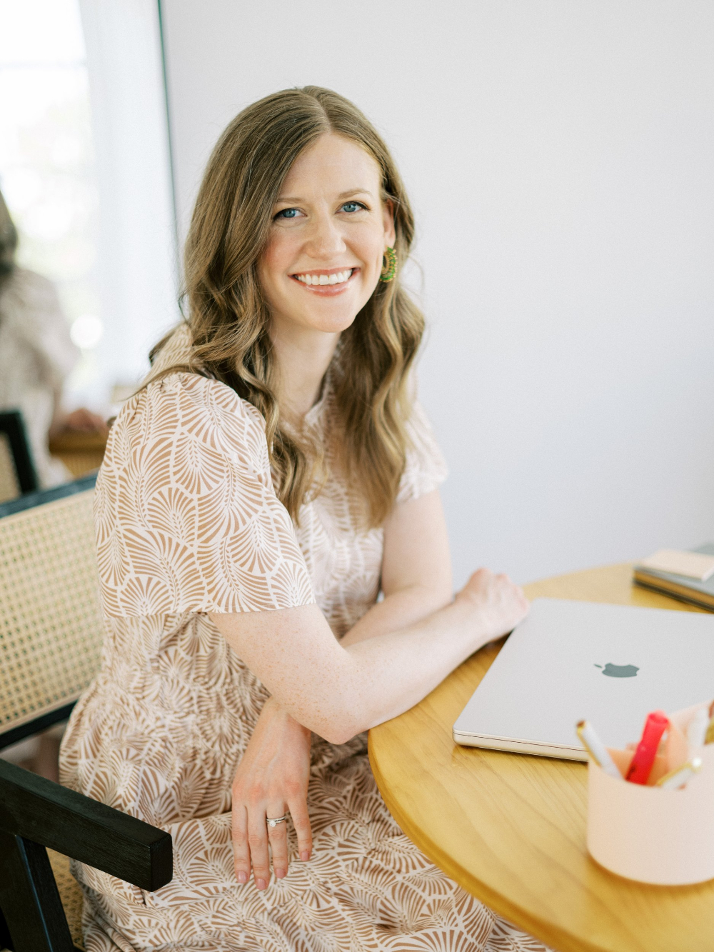 Smiling woman seated at a desk with a laptop, wearing a neutral patterned dress in a bright, modern workspace, representing a Showit web designer at work.