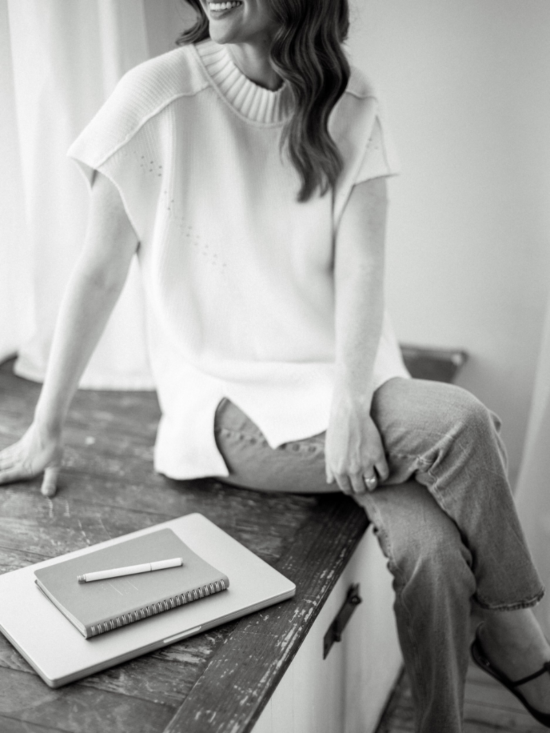 Woman seated on a wooden surface in a light top and jeans, relaxed posture in a softly lit space.
